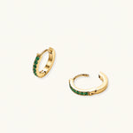 Load image into Gallery viewer, Pair of small gold huggie hoop earrings with bright green cubic zirconia stones on cream background