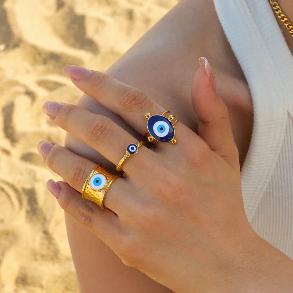 18k Bohemian Evil Eye Rings hand wearing two gold rings with blue evil eye designs against a blurred natural background.