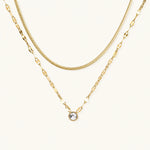 Load image into Gallery viewer, Layered gold necklace with herringbone and link chains, small bezel-set crystal pendant on cream background
