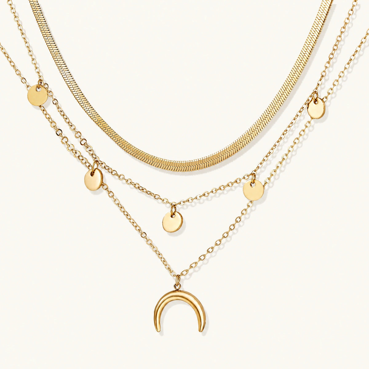 Four-layer gold necklace with herringbone chain, disc charms, and crescent moon pendant on cream background
