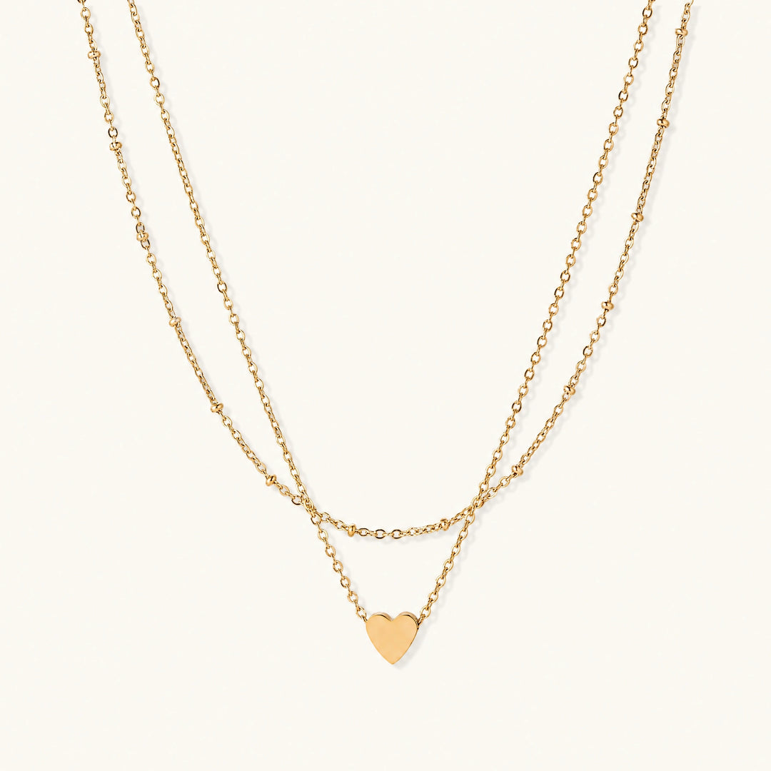 Delicate double-chain gold necklace with tiny heart pendant on cream background