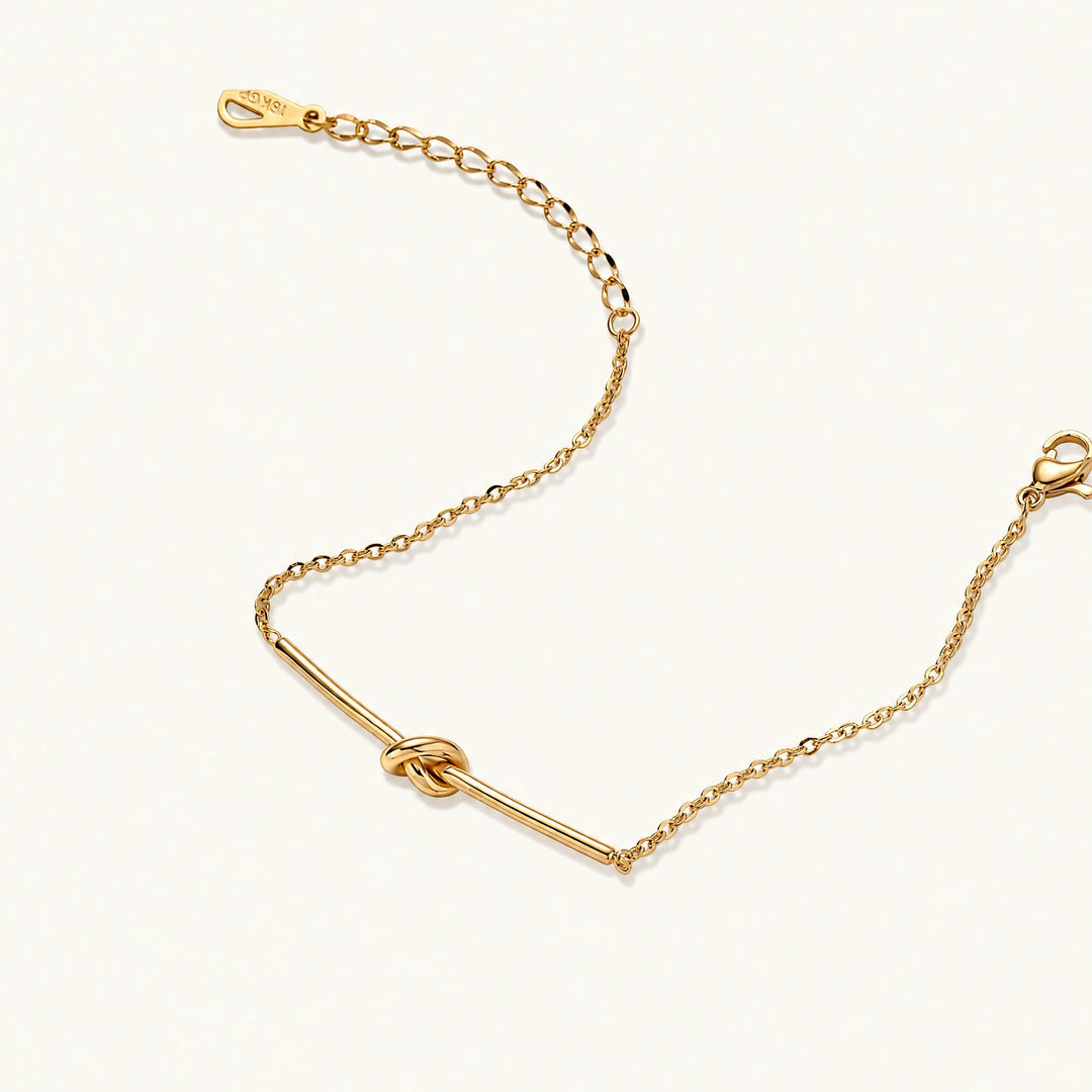 Delicate gold chain bracelet with minimalist bar and centered knot detail on cream background