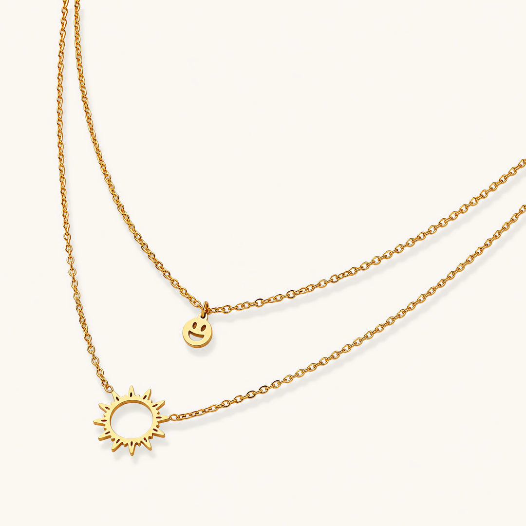 Gold layered necklace set with sunburst and smiley face pendants on delicate chains against white background.