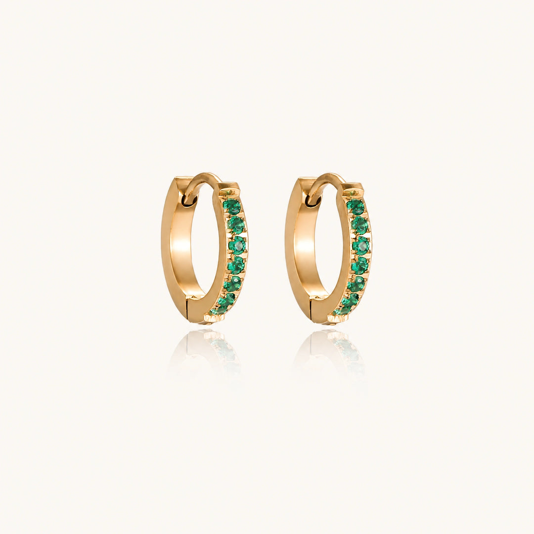 Pair of small gold huggie hoop earrings with bright green cubic zirconia stones on cream background