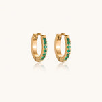 Load image into Gallery viewer, Pair of small gold huggie hoop earrings with bright green cubic zirconia stones on cream background
