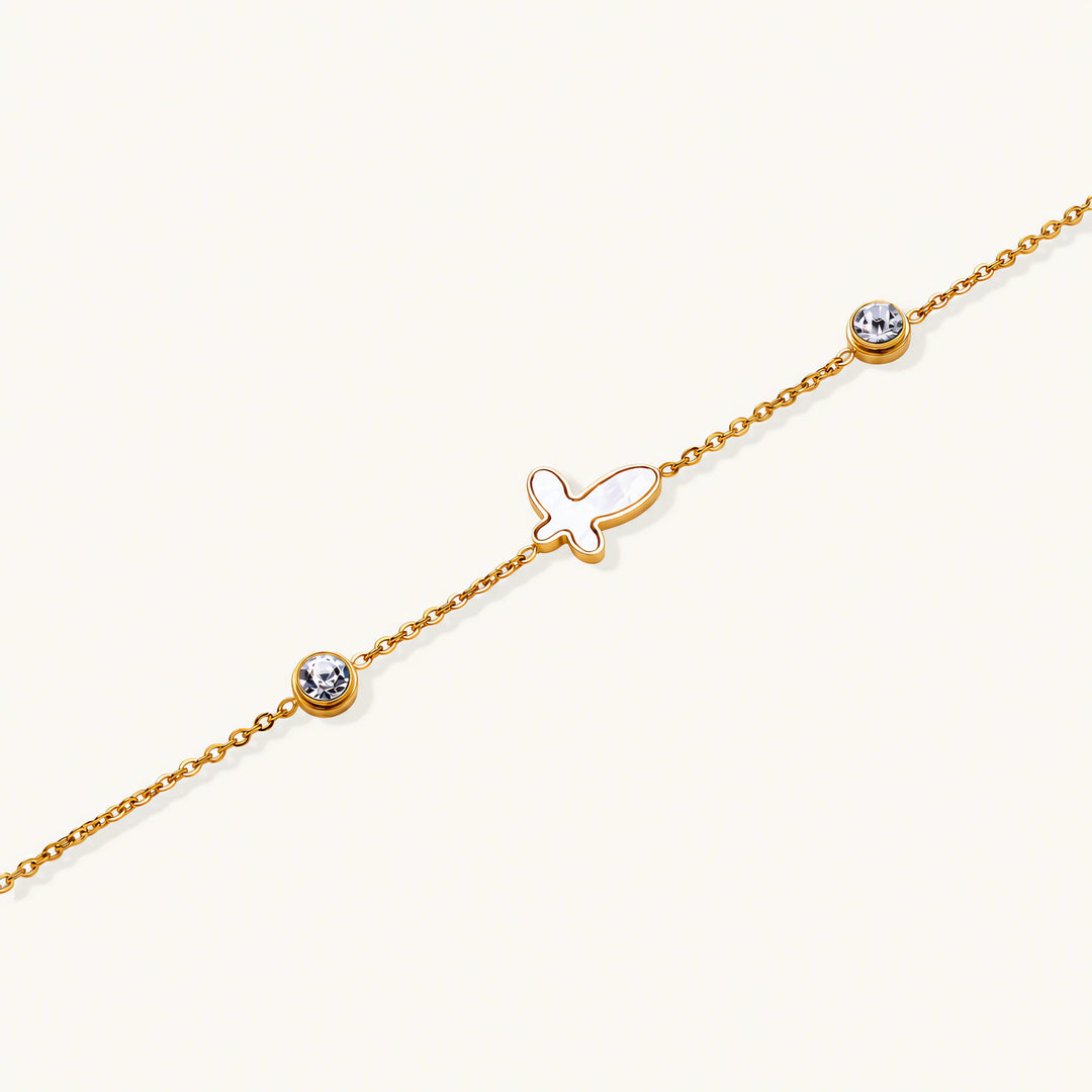 Gold chain bracelet with white mother of pearl butterfly pendant and two bezel-set crystals on cream background