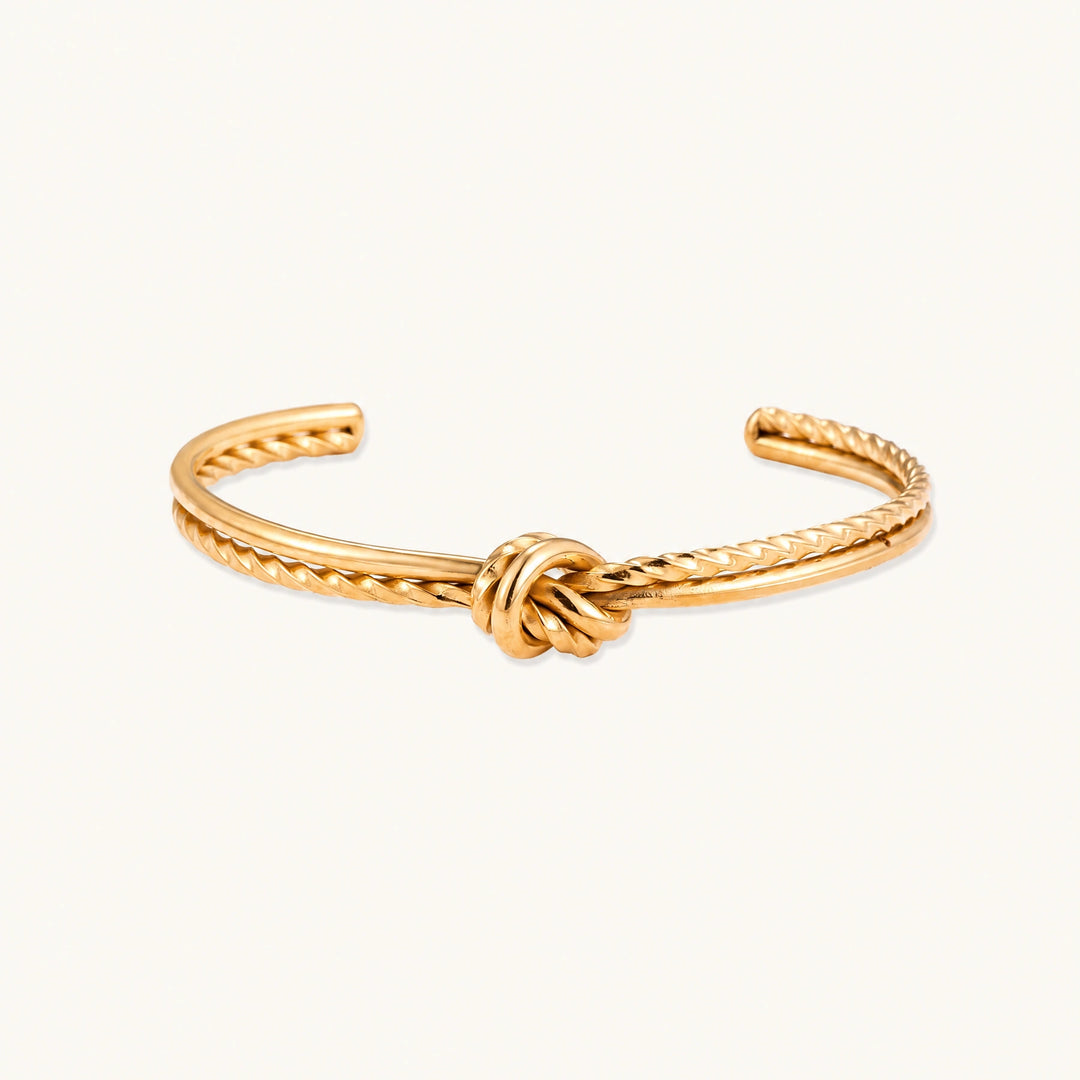 Gold cuff bracelet with twisted rope texture and decorative knot centerpiece on cream background.