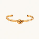 Load image into Gallery viewer, Gold cuff bracelet with twisted rope texture and decorative knot centerpiece on cream background.
