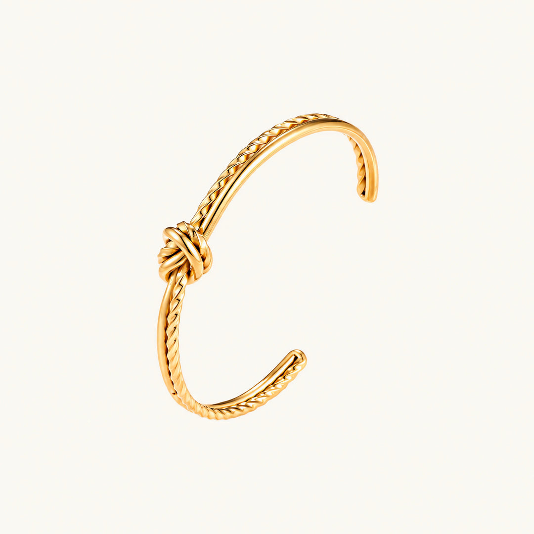 Gold cuff bracelet with twisted rope texture and decorative knot centerpiece on cream background.