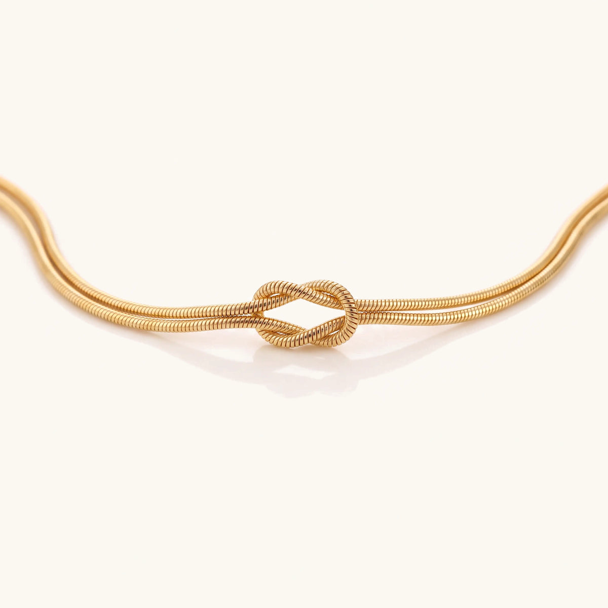 Fine double-strand gold snake chain bracelet tied in a reef knot on a cream background