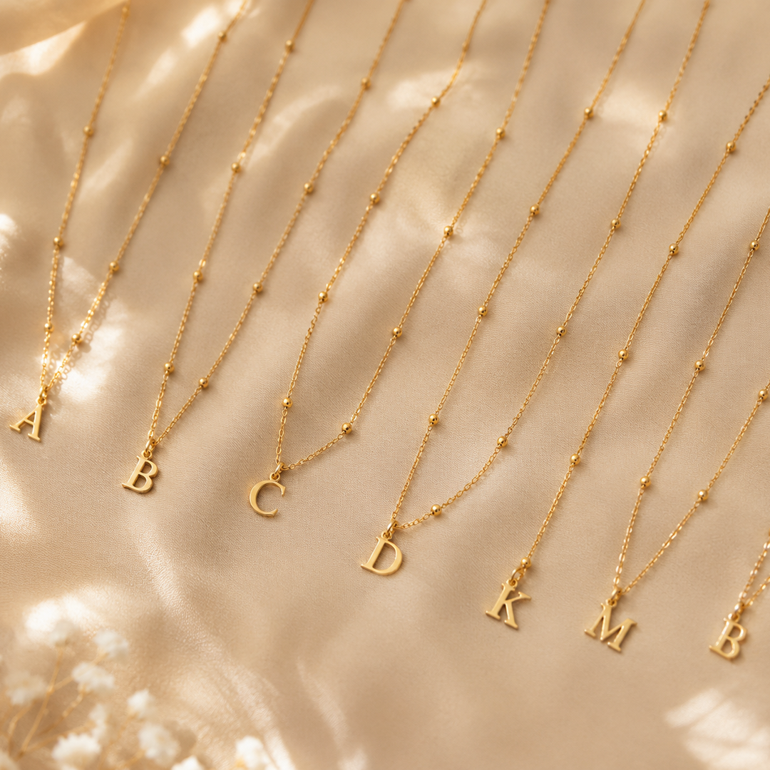 Dainty Satellite Chain Letter Initial Necklace delicate gold initial necklaces with A, B, C, D, K, M letter charms arranged on beige satin fabric.