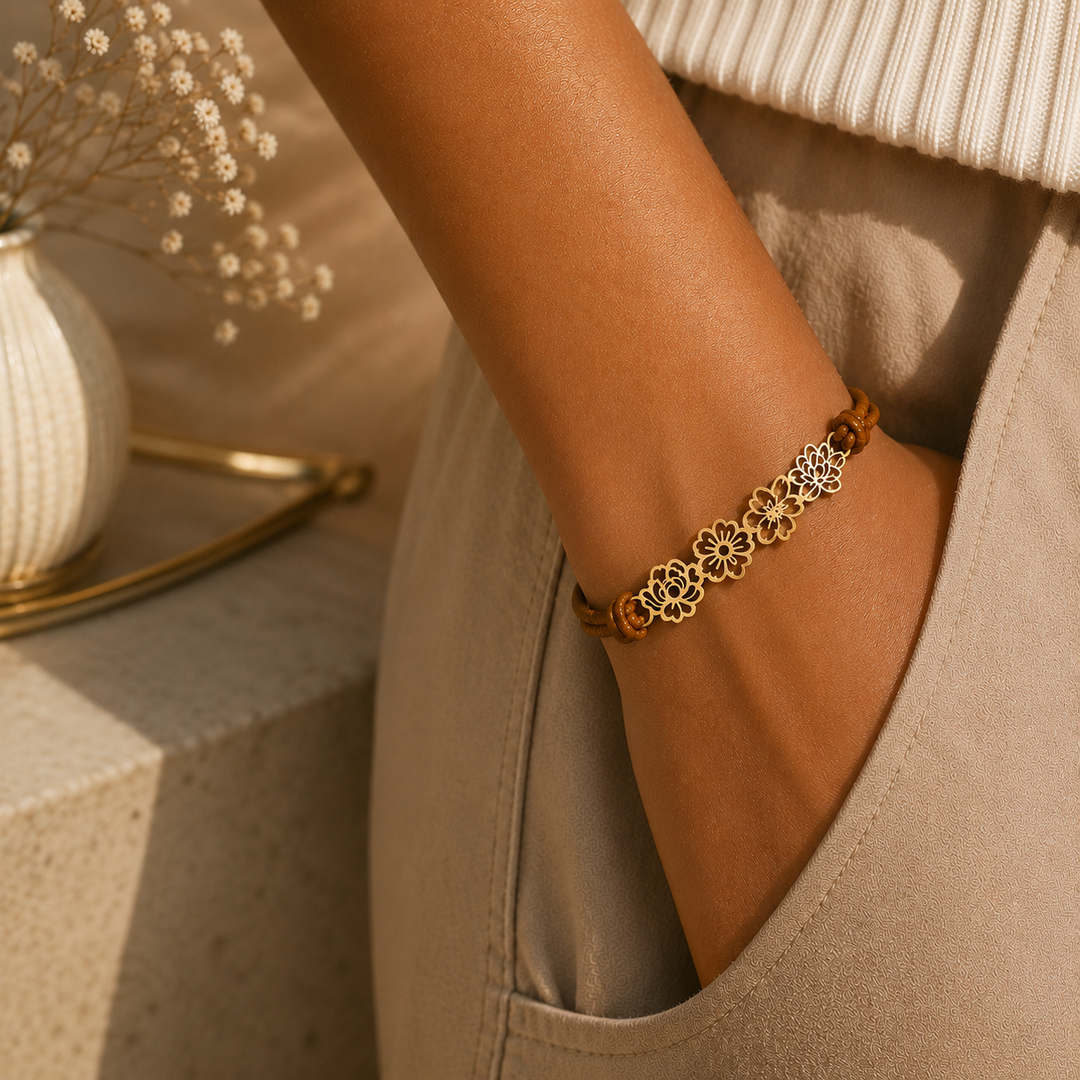 A close-up of a person's wrist wearing a delicate gold-tone bracelet with repeating floral motifs, paired with beige trousers and a cream ribbed top in warm natural light.