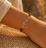 Load image into Gallery viewer, A delicate gold-tone floral bracelet with brown cord accents worn on a person's wrist, resting on a woven straw surface with a warm, sunlit background.
